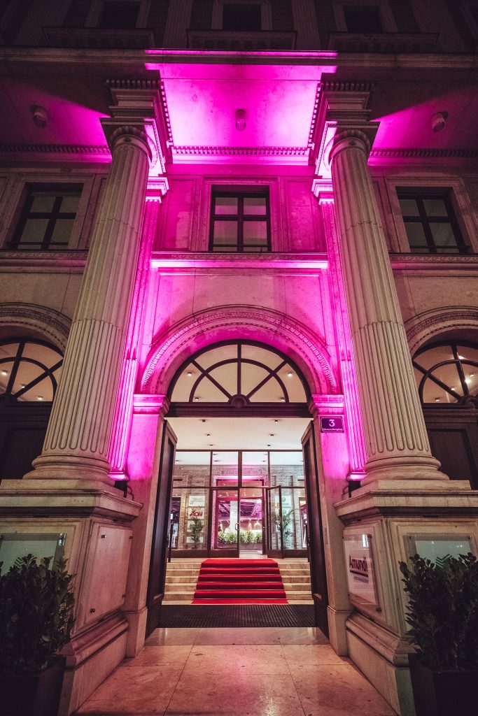 Neoclassical building entrance with two tall columns and pink nighttime lighting.