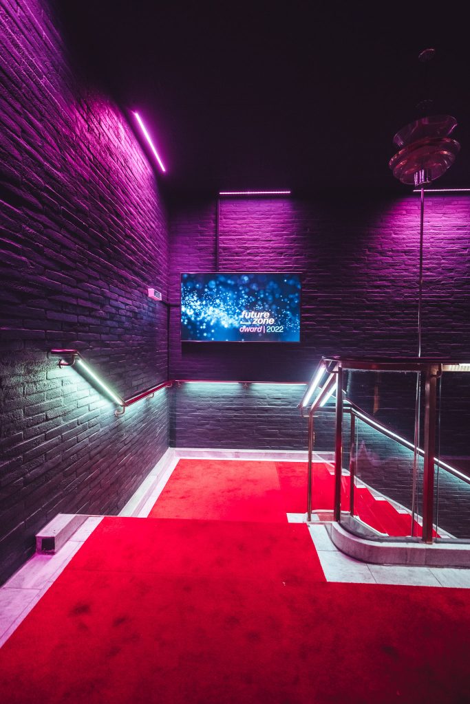 Stairwell with red carpet and brick walls bathed in purple neon light; wall screen shows 'future zone award 2022'.