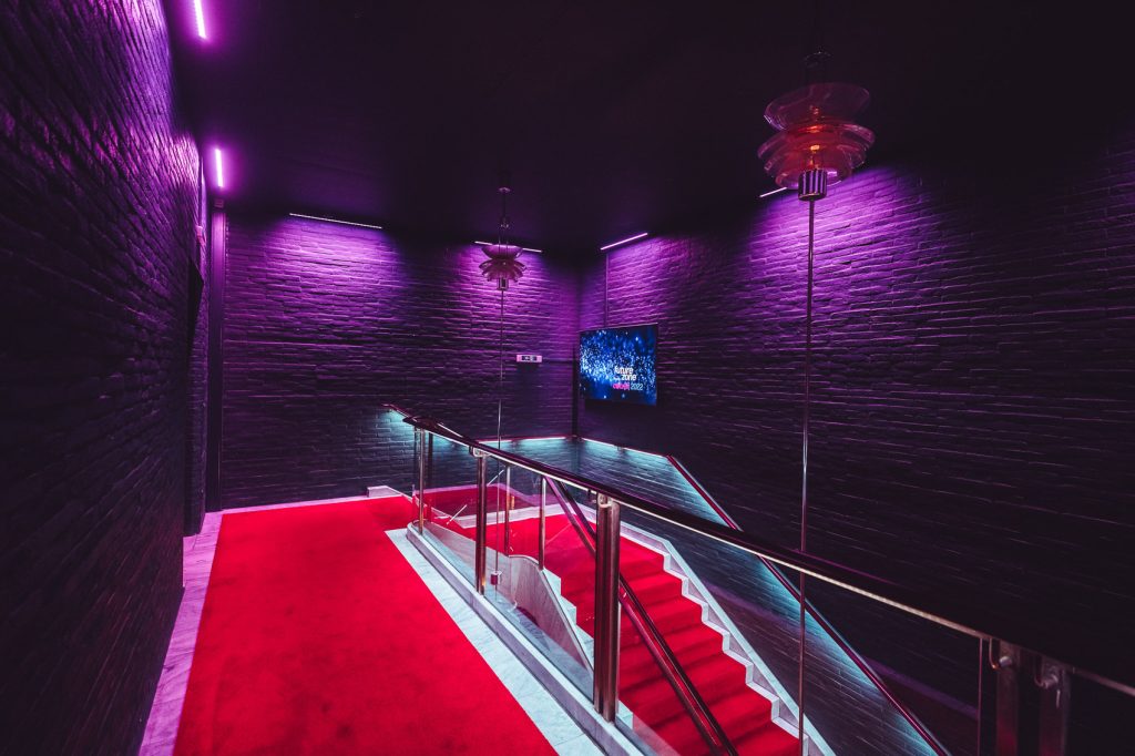 Dim neon-lit stairwell with red carpet and glass railing, purple ambient lighting on dark brick walls.