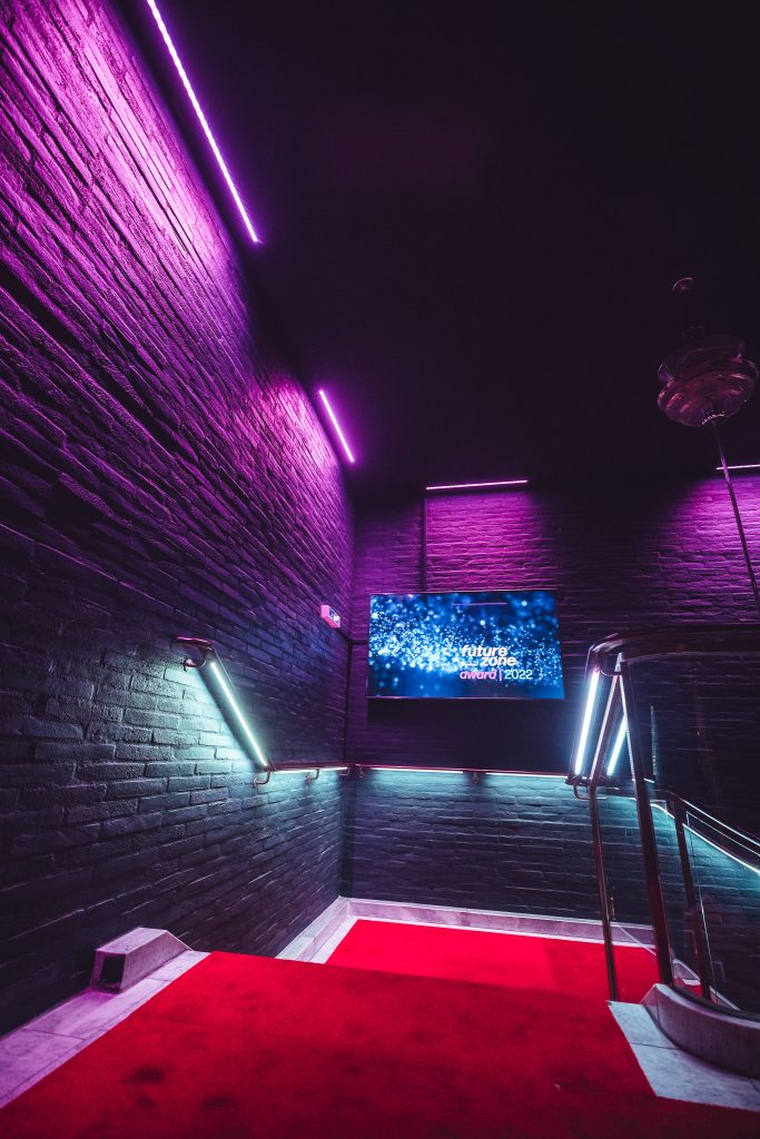 Dark brick stairwell bathed in purple and cyan neon, with a red carpet and a wall-mounted display screen.