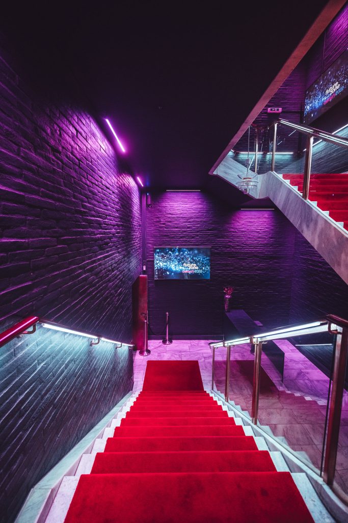 Top-down view of a red-carpet staircase descending a neon-lit, purple-hued brick-walled hallway with glass railing on the right.