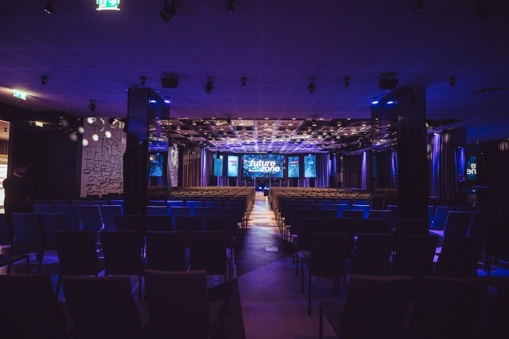 Dimly lit conference hall with rows of chairs facing a stage and LED screens showing 'future zone' at the front.