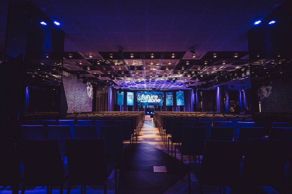 Dimly lit conference hall with rows of seats facing a lit stage and multiple screens displaying 'future zone' in blue hues.