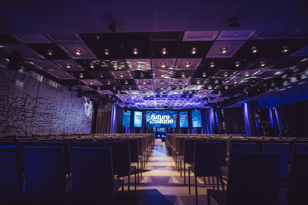 Empty conference hall with rows of chairs facing a lit stage; blue lighting and large screens display 'future zone' at the front.