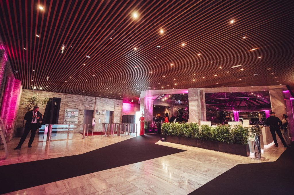 Wide hotel/event lobby with magenta ambient lighting, a black carpet runway, and guests near a reception area with greenery.