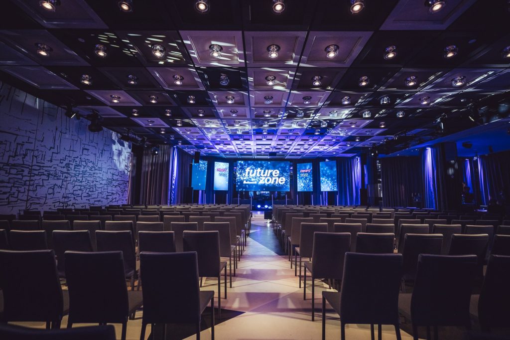 Empty conference hall with rows of chairs facing a blue-lit stage and large screens showing 'future zone'