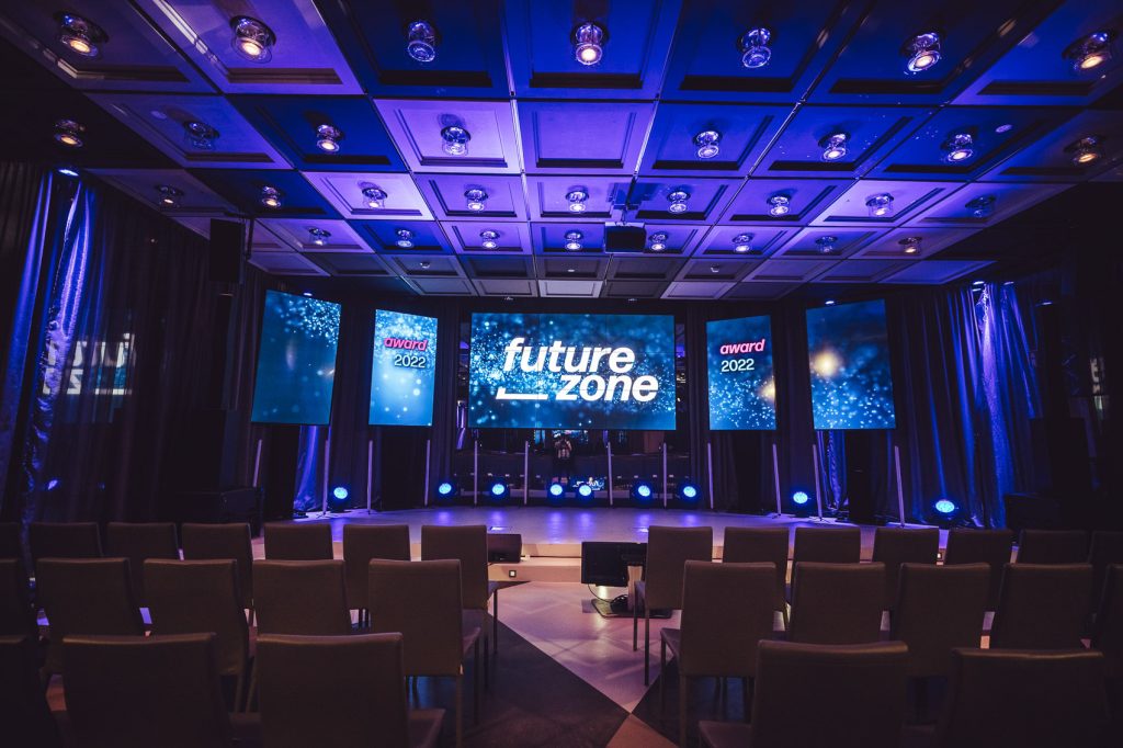 Dimly lit auditorium stage with blue lighting and four vertical screens displaying 'future zone' and 'award 2022' amid sparkly background, empty seats in front.