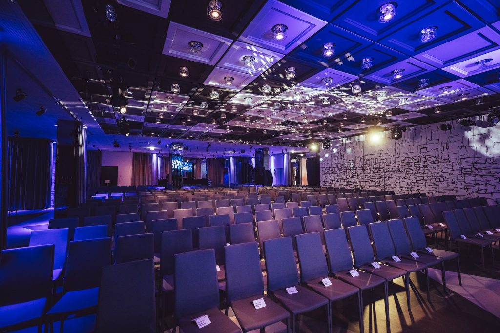 Large conference hall with rows of gray chairs facing a distant stage, bathed in blue and purple lighting.