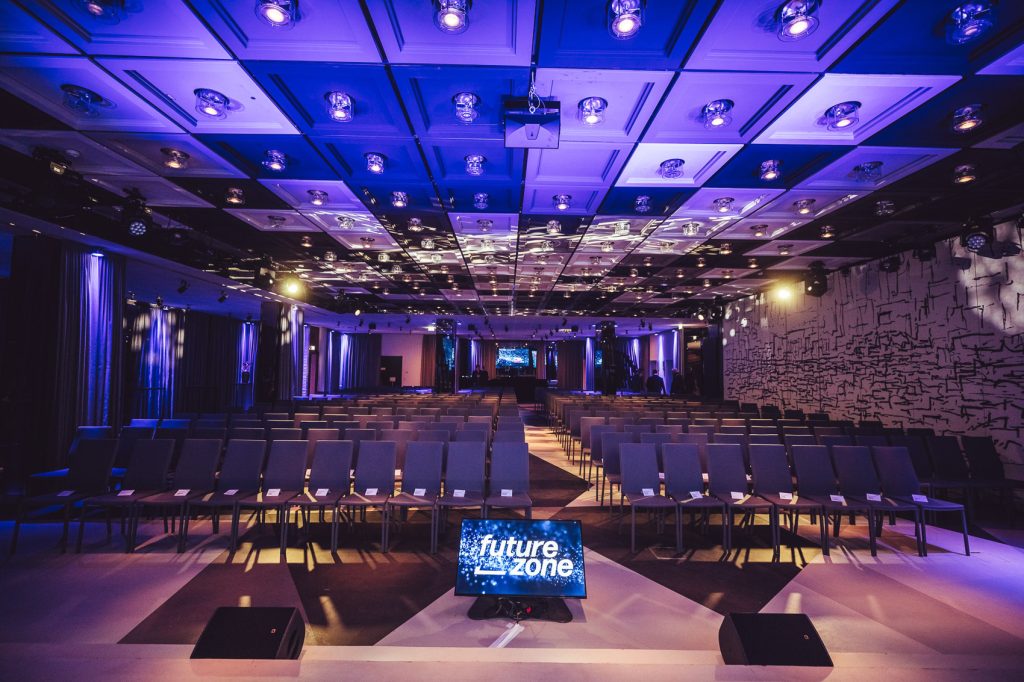 Conference hall prepared for a presentation with rows of chairs and a stage; screen shows 'future zone'.] }?Wait format. I must provide JSON with array. Let's craft properly.