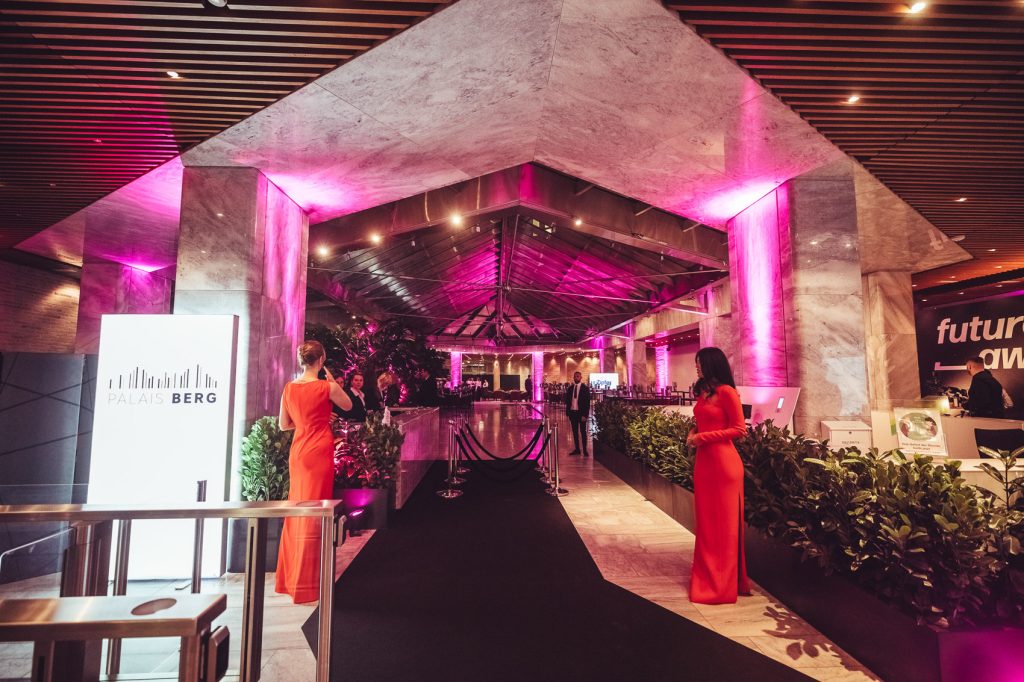 Event venue with pink uplighting and marble columns; a black carpet runway runs through the lobby, flanked by two women in red gowns near a Palais Berg sign.
