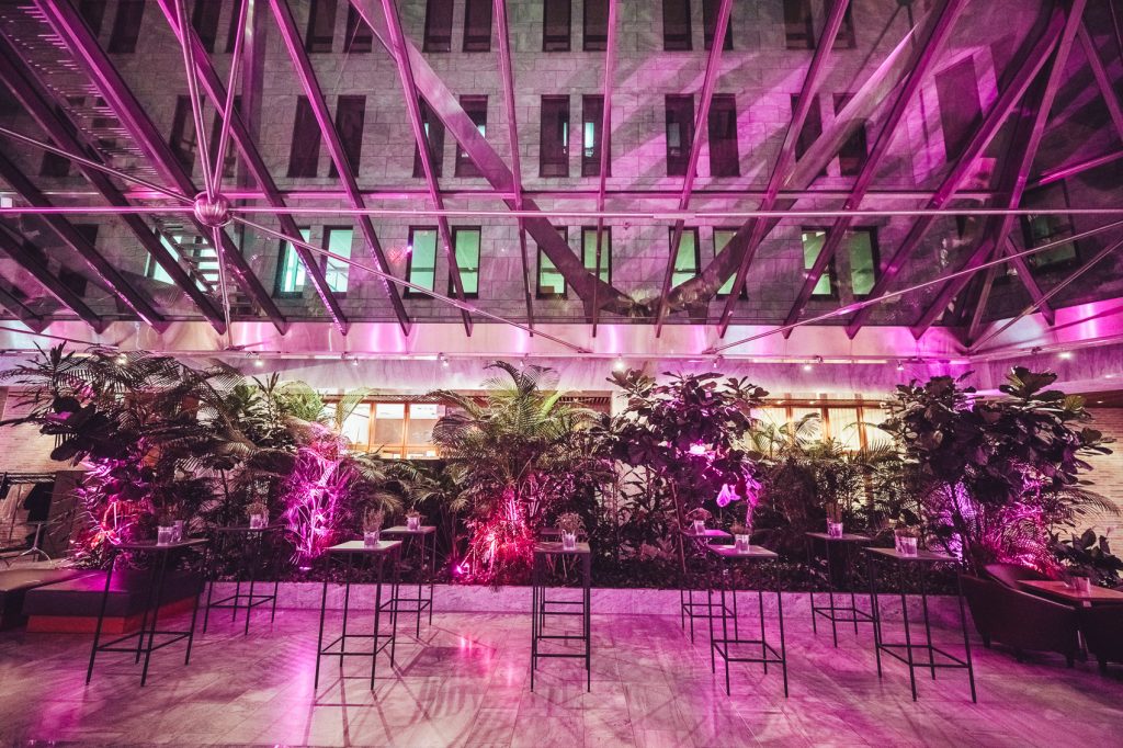 Indoor event space with pink-purple lighting, a row of tall tables and stools arranged in front of lush potted plants under a glass and steel geometric ceiling.