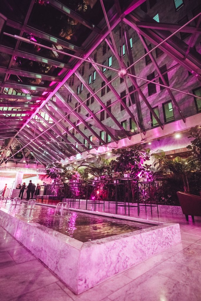 Indoor glass-roof atrium lit with pink lighting, shallow fountain along marble edge, and plants in the foreground with people in the background.