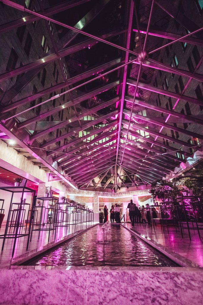 Indoor atrium with a glass, angular roof bathed in pink lighting and a central water feature with small fountains. People mingle near the back.