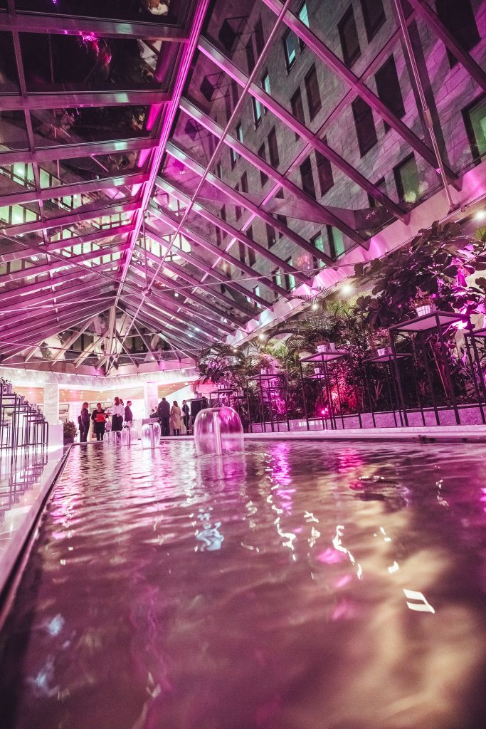 Atrium with a glass roof and pink neon lighting, a shallow pool in the foreground, and people mingling near a reception area.