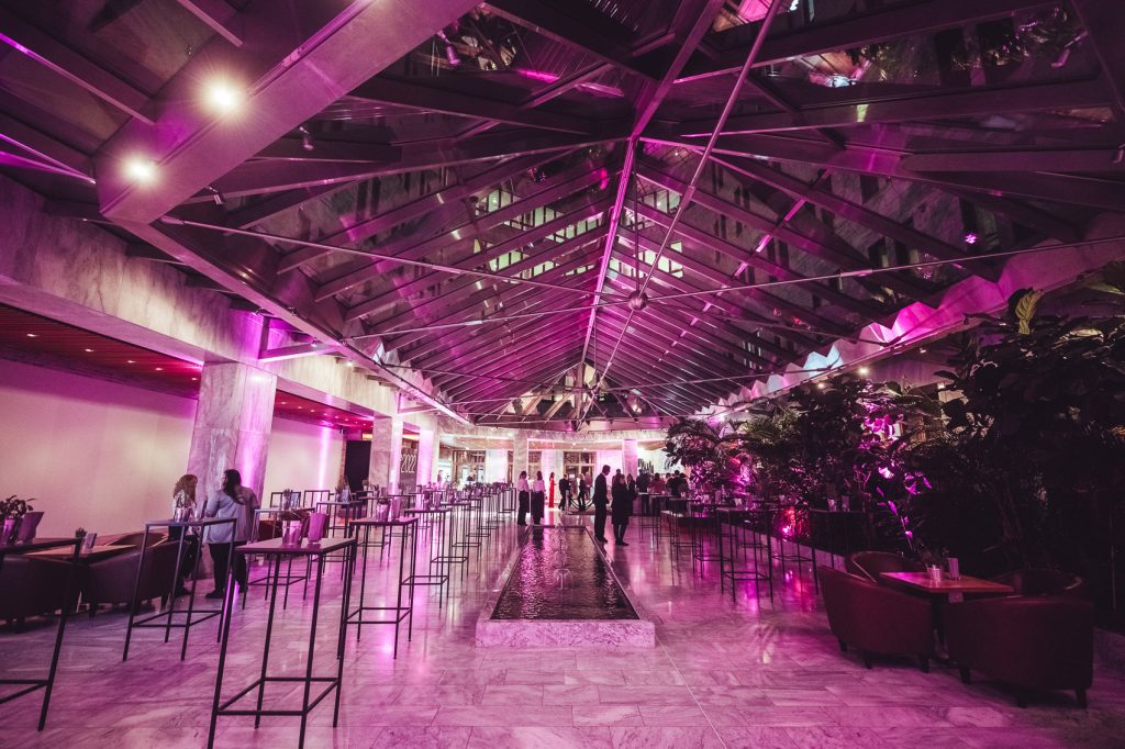 Indoor event hall with purple lighting, glass ceiling and metal beams, a central water feature, and guests mingling along a row of high tables.