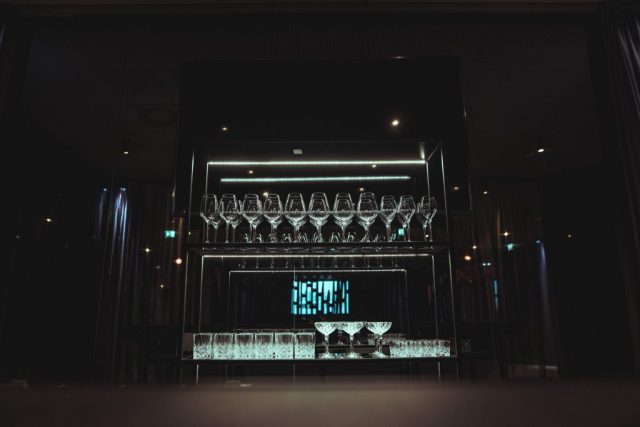 Rows of stemmed wine glasses hang upside down on a backlit glass shelf above a bar in a dim venue.