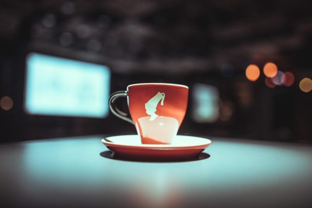 Red ceramic cup with white interior and a white design on the side sits on a matching saucer on a teal-lit surface, with blurred city lights in the background, creating bokeh.