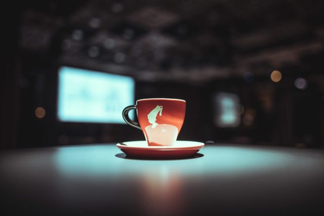 Red mug with a white graphic on a saucer, on a lit table with a dark, blurred background.