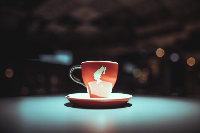 Red teacup with white design on a matching saucer, set on a teal table with blurred city lights in the background.