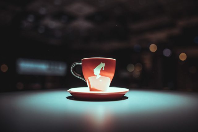 Red ceramic cup with a white graphic on a matching saucer, placed on a teal table with blurred city lights in the background.