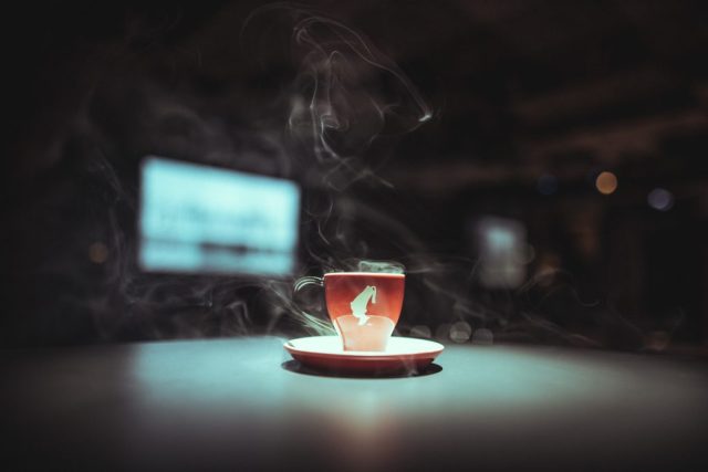 Red espresso cup on a matching saucer with curling steam against a dark, blurred background, suggesting a hot drink.
