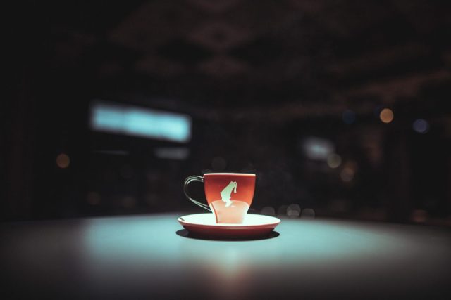 Red teacup with a white emblem on a matching saucer sits on a turquoise table, with blurred night lights in the background.