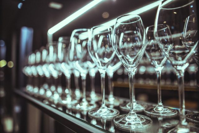 Row of empty wine glasses lined up on a bar shelf with blurred lights in the background.