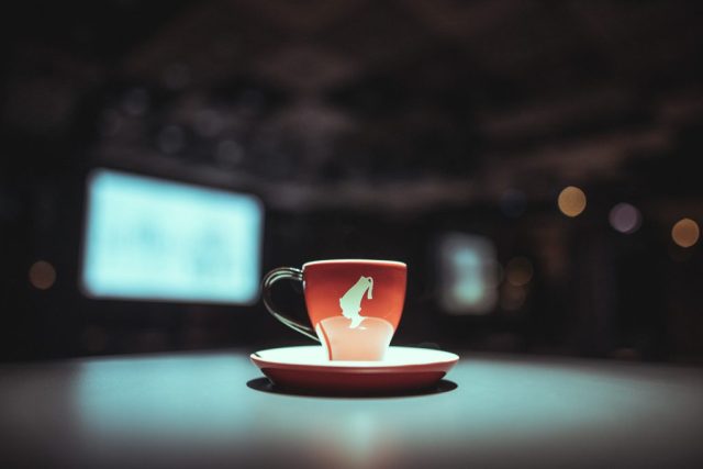 Red mug with a white horse logo on a matching saucer, set on a table with a blurred dark background and bokeh lights.