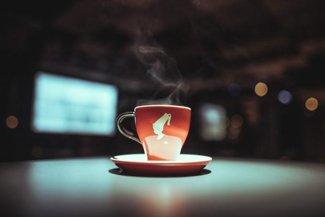 Red ceramic cup with steam rising on a matching saucer, set on a smooth surface with blurred city lights in the background.