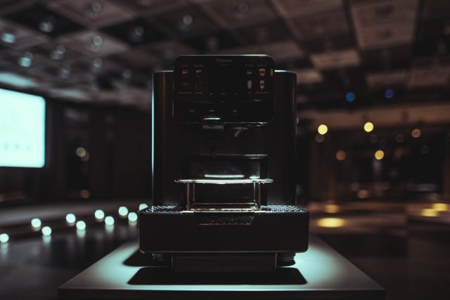 A dark-lit espresso machine on a counter in a cafe, with a glossy front and control panel visible.