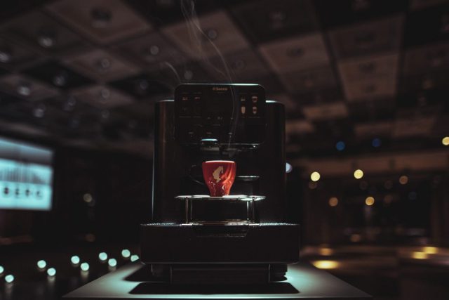 Close-up of a Saeco espresso machine with a red cup on the drip tray, steam rising in a dim cafe setting.