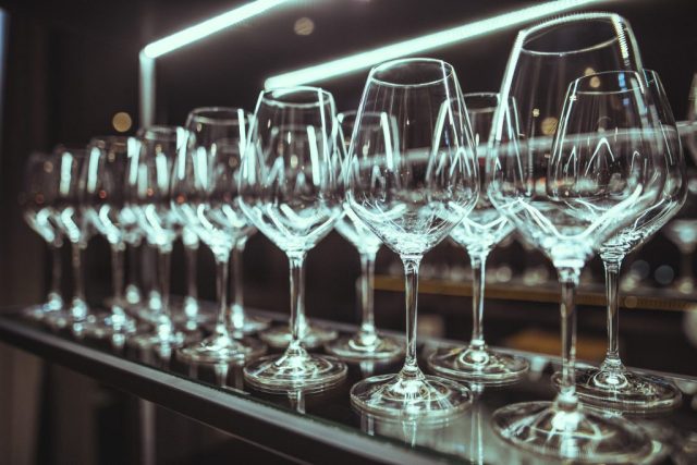 Row of wine glasses on a bar shelf with teal neon lighting and reflections in the glassware behind them