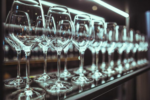 Row of wine glasses lined up on a mirrored bar surface with blue-tinted lighting and reflections.