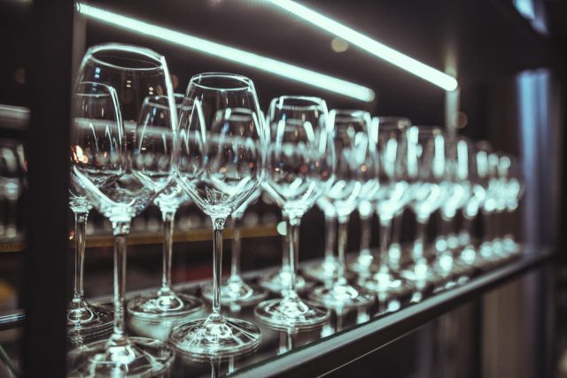 Row of wine glasses on a bar shelf with bright horizontal lights blurred in the background.
