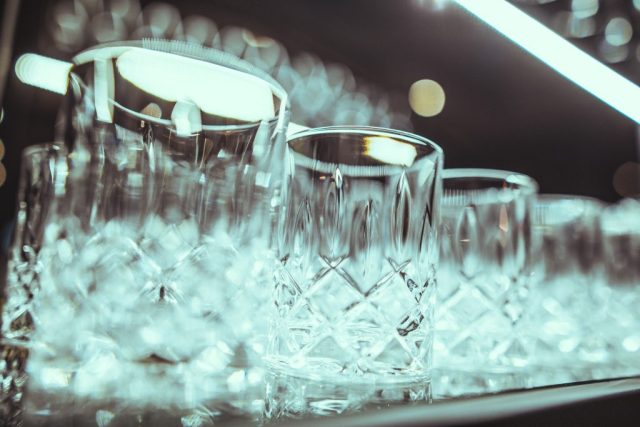 Row of crystal glasses with ice, reflections on the surface, soft bokeh lights in the background.