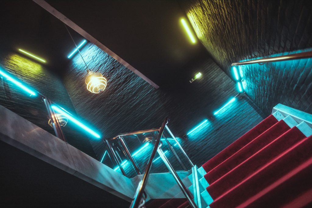 Angle view of a stairwell with red carpet, glass railing, and blue/green neon strips on dark brick walls, creating a neon-lit atmosphere.