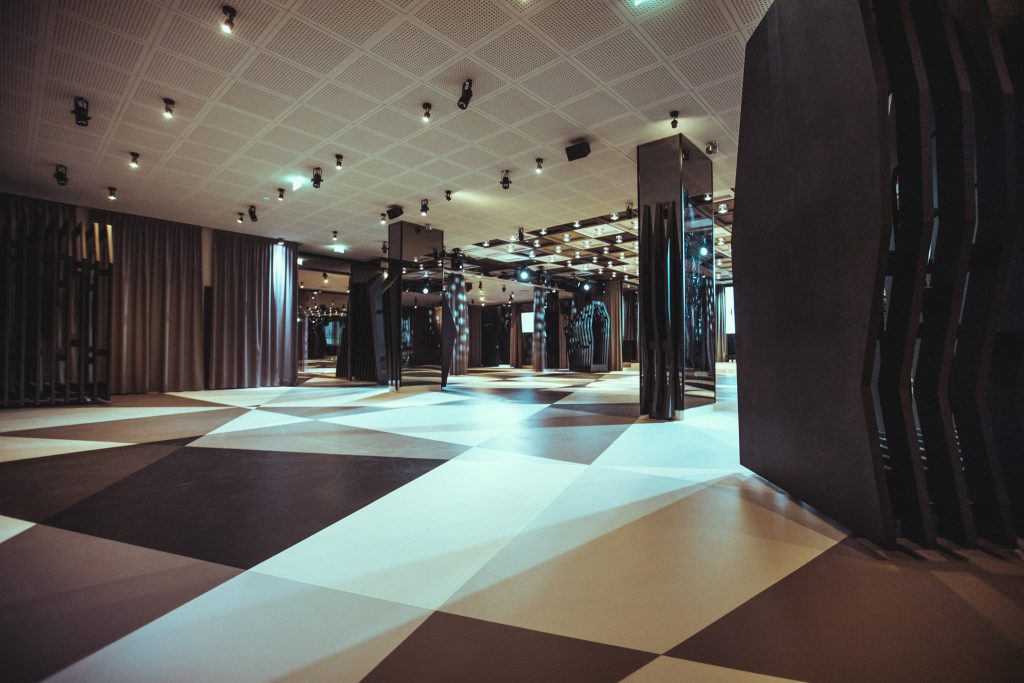Interior of a modern event hall with a black-and-white diamond-pattern floor, dark curtains, and tall black sculptural columns.