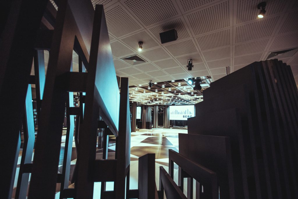 Modern conference hall interior with tall black geometric panels in foreground and a lit stage with screen in the background
