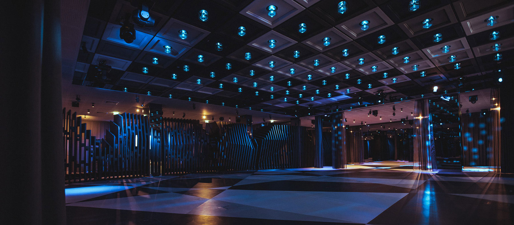 Dimly lit modern dance floor with blue lights, mirrored columns, and geometric wall panels.